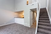 This two-story residence is defined by its striking soaring ceilings that create an expansive, airy atmosphere throughout the main living area. The open-concept lower level centers around a spacious carpeted living room that flows seamlessly into the kitchen via a breakfast bar with dark countertops. The kitchen is well-appointed with two-tone cabinetry—white uppers and gray lowers—complemented by a subway tile backsplash, dark stone countertops, and stainless steel appliances including a refrigerator and electric range. A white-railed staircase with a balcony overlook connects the main level to the upper floor, adding a distinct architectural focal point.

Upstairs, the bedrooms feature vaulted or elevated ceilings and plush carpeting. One bedroom benefits from generous natural light through large windows with vertical blinds, while another offers outdoor views through a standard window. A spacious walk-in closet with built-in shelving and hanging rods provides ample storage. The bathroom includes a full tub and shower combo with white tile surround, wood-look flooring, a vanity with dark countertops, and neutral gray walls. Throughout, the neutral color palette and soft carpeting contribute to a clean, cohesive ambiance.
