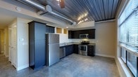 This single-floor space showcases a striking industrial aesthetic that seamlessly blends raw architectural elements with modern finishes. The open-concept kitchen serves as the centerpiece, outfitted with a full suite of stainless steel appliances including a French door refrigerator, electric range, built-in microwave, and dishwasher. Dark espresso cabinetry with granite-look countertops provides a bold contrast against the neutral walls, offering ample storage and a clean, contemporary look. The polished concrete floors extend throughout the space, reinforcing the industrial character while remaining easy to maintain. Above, exposed silver ductwork runs along a corrugated metal ceiling, lending an urban, loft-like ambiance to the interior. Track lighting mounted along the ceiling provides focused illumination throughout the kitchen and open living area. The open layout creates a generous sense of space and flow, making the area feel expansive and versatile. The combination of raw materials, modern appliances, and thoughtful design details gives this space a distinctive personality well-suited to contemporary urban living.