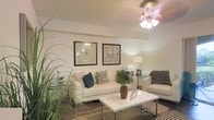 This single-floor space offers a thoughtfully designed interior that balances comfort and style. The living area serves as the focal point of the home, showcasing warm, neutral-toned walls that create a welcoming and serene atmosphere. A textured ceiling adds subtle architectural character overhead, while dark hardwood-style flooring grounds the space with a rich, classic contrast.

The living room is furnished with a crisp white sofa accented by decorative teal and patterned throw pillows, complemented by a grey upholstered chair. A sleek white coffee table and a glass side table add modern touches, while a ceiling fan with an ornate light fixture provides both function and a decorative element.

Two large framed botanical art pieces are symmetrically displayed on the main wall, contributing to the space's cohesive and curated aesthetic. Lush indoor plants are thoughtfully placed throughout, bringing natural vitality and a refreshing organic feel to the interior. A floor lamp adds warm ambient lighting, enhancing the inviting quality of the room.

Overall, this space presents a clean, contemporary living environment with carefully chosen furnishings and décor that create a harmonious and comfortable atmosphere throughout.