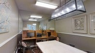 This single-floor workspace is designed to accommodate both focused individual work and collaborative activity. The room features a row of built-in wooden cubicle-style workstations along one wall, each equipped with a desktop computer monitor and individual desk lamps, providing semi-private areas well-suited for personal tasks or computer work. A printer station is conveniently integrated into the workstation area, adding practical functionality to the setup. Overhead fluorescent lighting ensures the space is well-lit throughout, supplemented by a distinctive multi-bulb pendant light fixture that adds an industrial accent to the ceiling. A large communal table with metal chairs occupies the central area, offering additional surface space for group work or casual use. The neutral wall tones and textured ceiling contribute to a calm, utilitarian atmosphere. Natural light filters in through glass-paneled doors visible at the far end of the room, providing a connection to the exterior. The overall layout is practical and efficiently organized, making the space a functional environment for work, study, or resource use.