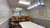 This single-floor workspace is designed to accommodate both focused individual work and collaborative activity. The room features a row of built-in wooden cubicle-style workstations along one wall, each equipped with a desktop computer monitor and individual desk lamps, providing semi-private areas well-suited for personal tasks or computer work. A printer station is conveniently integrated into the workstation area, adding practical functionality to the setup. Overhead fluorescent lighting ensures the space is well-lit throughout, supplemented by a distinctive multi-bulb pendant light fixture that adds an industrial accent to the ceiling. A large communal table with metal chairs occupies the central area, offering additional surface space for group work or casual use. The neutral wall tones and textured ceiling contribute to a calm, utilitarian atmosphere. Natural light filters in through glass-paneled doors visible at the far end of the room, providing a connection to the exterior. The overall layout is practical and efficiently organized, making the space a functional environment for work, study, or resource use.