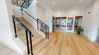 Located at 7207 McKnight Rd in Pittsburgh, PA, this contemporary amenity space serves as a welcoming lobby and fitness center entrance for its building. The space is defined by its clean, modern aesthetic, showcasing wide-plank light hardwood flooring that runs seamlessly throughout and crisp white walls that amplify the sense of openness.

The main lobby area features a sleek reception desk with warm leather seating, a wall-mounted display screen, and a corridor leading to additional rooms. Large floor-to-ceiling glass entry doors flood the space with natural light, offering views to the exterior and creating an inviting, airy atmosphere. A branded Founding House welcome mat marks the main entrance.

A striking staircase with light wood treads and a modern black metal and glass railing serves as a central architectural feature, ascending to an upper-level mezzanine with glass balustrade overlooking the lobby below. Recessed lighting and elevated ceilings throughout contribute to the bright, polished ambiance.

Adjacent to the lobby, a fully equipped fitness center is accessible through glass-paneled doors with bold red accents, offering visibility into the workout area stocked with exercise equipment. A decorative potted plant adds a natural touch near the gym entrance. Overall, the space presents a highly functional yet stylish environment tailored for modern communal living.