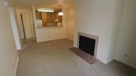 This single-floor residence offers a practical and comfortable layout designed for everyday living. The main living area is carpeted throughout, providing a warm and inviting atmosphere. A standout feature of this space is the tile-surround fireplace with a classic brick hearth, serving as a natural focal point of the living room and adding a cozy, welcoming character to the area.

The kitchen is open to the living space via a pass-through counter that doubles as a breakfast bar, enhancing the sense of connectivity between the two areas. Warm, medium-toned wood cabinetry lines the kitchen, complemented by a white refrigerator and under-cabinet fluorescent lighting that ensures a well-lit workspace. A pendant light hangs above the pass-through counter, adding a touch of character to the kitchen area.

The neutral color palette throughout — from the beige carpeting to the white walls — creates a clean, versatile backdrop that adapts easily to various furnishing styles. The overall layout is straightforward and efficient, making good use of the available space. The open sightlines between the kitchen and living area contribute to an airy, connected feel throughout the home.