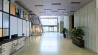 This contemporary commercial space presents a striking first impression through its expansive, open-plan lobby. The highly polished concrete floors reflect natural light that pours in through a dramatic floor-to-ceiling glass façade, lending the space an airy, luminous quality. A sleek open-tread staircase with a dark metal frame serves as both a functional and architectural focal point, rising elegantly against the backdrop of the glazed exterior wall.

The ceiling features a structured grid of recessed and drop panels outfitted with integrated lighting, providing even illumination throughout the space. On one side, a glass-partitioned area offers a glimpse into an adjacent workspace or retail zone, adding depth and transparency to the overall layout. Textured stone or concrete columns punctuate the right-hand side of the lobby, contributing an industrial yet refined aesthetic.

Strategically placed large planters with lush fiddle-leaf fig trees introduce a natural, organic element, softening the hard surfaces and adding warmth to the environment. A reception or service counter is positioned to one side, completing the functional arrangement of the space. The overall design balances industrial materials with modern finishes, creating a professional, welcoming, and visually dynamic environment suited for a corporate or mixed-use setting.