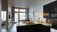 This single-floor space showcases a well-designed open-concept layout that seamlessly connects the kitchen, dining, and living areas into one cohesive living environment. The kitchen is a standout feature, equipped with dark cabinetry, a double-basin sink, and granite-look countertops that extend into a peninsula, offering both prep space and a casual dining surface. Pendant lighting with warm amber shades hangs above, adding a refined yet inviting ambiance to the kitchen zone.

The living and dining areas benefit from generous natural light streaming through large floor-to-ceiling windows and a glass sliding door that leads to a balcony, providing views of the surrounding exterior. Bold geometric-patterned curtains frame the windows, adding a decorative contrast to the neutral gray walls and light wood-style flooring that runs throughout the space.

The overall aesthetic is modern and cohesive, combining dark and light tones with carefully chosen accent pieces. Wall art and tasteful decor elements contribute to an inviting atmosphere, while the open layout ensures a sense of spaciousness and ease of flow between the functional zones.