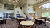 This expansive common area is defined by its striking double-height ceilings, which lend an open, airy quality to the space. Two symmetrical staircases with light wood railings flank the room, leading to an upper-level mezzanine or loft area, adding architectural interest and vertical dimension. Recessed lighting is complemented by a decorative chandelier, ensuring the space is well-lit throughout.

The main floor is thoughtfully arranged to serve multiple functions. A dining area features several round tables paired with dark wood chairs, positioned atop a richly patterned carpet that anchors the space. Toward the rear, a comfortable lounge area includes upholstered seating arranged around a television and a fireplace, providing a relaxed setting for socializing. Decorative wall art, including a bird motif and floral arrangements, adds warmth and character to the neutral-toned walls.

Natural light filters in through windows and a glass door, connecting the interior to the outdoors. Overall, the space balances functionality with a welcoming ambiance suited for communal gatherings and everyday activities.