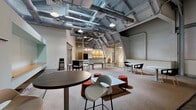 Located at 120 Perry St in Petersburg, VA, this single-floor commercial space offers a generous open-plan layout with a striking industrial aesthetic. The most commanding architectural feature is the dramatically high ceiling, which showcases exposed steel structural arches, corrugated metal panels, and large-diameter HVAC ductwork — all lending the space a raw, urban character. Pendant lighting and track fixtures hang from the ceiling framework, providing layered illumination throughout.

The floor transitions between polished concrete and carpeted areas, effectively delineating distinct functional zones within the open layout. A carpeted central gathering area accommodates a variety of modern furnishings, including round tables with contemporary seating and modular stools, making it well-suited for collaborative work, meetings, or social events. A second open zone with exposed concrete flooring provides a more flexible, multipurpose area.

A built-in kitchenette features warm wood cabinetry, granite-style countertops, a double sink, and ample counter space. Nearby, large industrial-style double doors provide access to an adjacent utility or storage area. Exposed brick and painted masonry walls add texture and reinforce the space's industrial character. The overall ambiance is modern and versatile, balancing raw architectural elements with thoughtfully arranged collaborative zones.