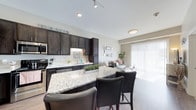 This single-floor residence offers a thoughtfully designed open-concept layout that seamlessly connects the kitchen and living areas, creating an inviting and functional living space. The kitchen is a standout feature, appointed with rich dark cabinetry that extends to the ceiling, providing ample storage. A large granite-topped island with seating for two serves as the centerpiece, ideal for casual dining or entertaining. Stainless steel appliances, including a range, dishwasher, and microwave, complement the sleek aesthetic, while under-cabinet lighting adds a warm, practical touch. Light-toned countertops and a clean white backsplash brighten the space and contrast elegantly with the dark cabinetry. The kitchen flows naturally into the living area, which is bathed in natural light streaming through large sliding glass doors fitted with vertical blinds. The living space is open and airy, with neutral-toned walls and light hardwood-style flooring that run throughout, lending cohesion to the overall design. Track lighting and recessed ceiling fixtures ensure the space is well-lit at all times. The combination of modern finishes, functional design, and an abundance of natural light makes this residence a comfortable and stylish place to call home.