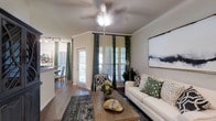 This single-floor residence offers a thoughtfully designed interior that balances comfort and style. The living area serves as the heart of the space, showcasing rich hardwood flooring, crisp crown molding, and a ceiling fan that adds both function and a classic touch overhead. Large windows dressed with bold green patterned drapes allow generous natural light to fill the room, creating a warm and inviting ambiance throughout the day. The living space is furnished with a clean white sofa accented by decorative pillows in coordinating green tones, centered around a wooden coffee table atop a layered area rug. A patterned accent chair adds visual interest and depth to the seating arrangement. A framed piece of abstract wall art enhances the contemporary aesthetic of the room. Just beyond the living area, a glimpse into the dining space reveals additional hardwood flooring, a dining set, and track lighting overhead, suggesting a seamless and open flow between living and dining areas. A glass-paneled door connects the interior to an exterior access point, further brightening the space. The overall atmosphere is elegant yet approachable, with a cohesive color palette of greens, whites, and warm wood tones tying each area together beautifully.