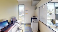 This university business center computer room is a compact, single-floor space designed to support residents with essential productivity needs. The room features multiple desktop workstations arranged along warm wood-toned desks paired with ergonomic white rolling chairs, providing a functional and comfortable setup for computing tasks. A large multifunction printer and copier station is conveniently positioned near the entrance, making printing and scanning easily accessible. The walls are accented with a bold yellow-green tone on one side, adding a vibrant, modern character to the space, while the remaining walls are finished in clean white. A tall, narrow window along one wall invites ample natural light and offers views of the landscaped exterior grounds. The flooring consists of a patterned carpet tile in a mix of blue, yellow, and grey tones, contributing to the room's contemporary aesthetic. A posted notice displays the business center rules, reflecting the organized and community-oriented nature of the facility. A glass-panel door connects the computer room to an adjacent recreation lounge, where a pool table is visible, highlighting the room's integration within a broader amenity hub. The overall atmosphere is bright, functional, and purposefully designed for academic and professional use.