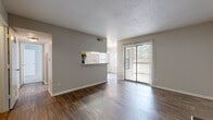 This single-story unit is thoughtfully laid out to maximize its compact footprint, combining the living, dining, and kitchen areas into one cohesive open-plan space. The living room is anchored by a black stone fireplace and a sliding glass door fitted with vertical blinds, which fills the room with natural light and provides direct access to an outdoor area. Dark hardwood-style flooring runs throughout the main living spaces, adding warmth and visual continuity.

The kitchen features warm oak cabinetry, a mosaic tile backsplash, black appliances including a range and refrigerator, and granite-style countertops. A pass-through window with a breakfast bar counter connects the kitchen to the living area, enhancing the open feel of the space.

A hallway leads from the main living area to the private bedroom, which is carpeted and features a large window with a view of greenery, providing ample natural light. Two closets offer practical storage throughout the unit.

The bathroom includes a full tub-shower combination with white tile surround, a wood-toned vanity with a granite-style countertop, and hardwood-style flooring that mirrors the rest of the unit. The overall palette of neutral grays and warm wood tones creates a clean, cohesive aesthetic throughout.