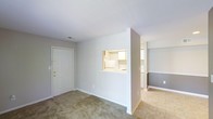 This single-floor residence offers a thoughtfully designed layout that maximizes both functionality and flow. The living area greets you upon entry through a solid white front door with gold-toned hardware, setting a clean and welcoming tone. Soft, neutral-colored carpet covers the living space floor, adding warmth underfoot, while the walls are finished in a light gray palette that lends a calm, understated ambiance throughout.

A defining architectural feature is the pass-through window cut into the dividing wall between the living area and kitchen, offering convenient sight lines and an open, connected feel without fully removing the separation between spaces. The kitchen is outfitted with white cabinetry and appliances, maintaining the consistent neutral color scheme.

Beyond the kitchen partition, the space transitions into a hallway or adjoining room with two-tone walls — a lighter upper half and a deeper gray lower half — adding a subtle design detail that gives depth to the interior. The flooring in this area shifts to light-toned tile, creating a natural visual distinction between zones.

The layout flows efficiently across its single level, making the space easy to navigate. The overall design is clean, minimal, and move-in ready, suited for those who appreciate a simple yet practical living environment.