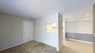 This single-floor residence offers a thoughtfully designed layout that maximizes both functionality and flow. The living area greets you upon entry through a solid white front door with gold-toned hardware, setting a clean and welcoming tone. Soft, neutral-colored carpet covers the living space floor, adding warmth underfoot, while the walls are finished in a light gray palette that lends a calm, understated ambiance throughout.

A defining architectural feature is the pass-through window cut into the dividing wall between the living area and kitchen, offering convenient sight lines and an open, connected feel without fully removing the separation between spaces. The kitchen is outfitted with white cabinetry and appliances, maintaining the consistent neutral color scheme.

Beyond the kitchen partition, the space transitions into a hallway or adjoining room with two-tone walls — a lighter upper half and a deeper gray lower half — adding a subtle design detail that gives depth to the interior. The flooring in this area shifts to light-toned tile, creating a natural visual distinction between zones.

The layout flows efficiently across its single level, making the space easy to navigate. The overall design is clean, minimal, and move-in ready, suited for those who appreciate a simple yet practical living environment.