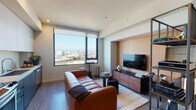 Located at 11200 W Pico Blvd in Los Angeles, this thoughtfully designed unit occupies an elevated floor, offering sweeping views of the Los Angeles cityscape through large, black-framed windows that flood the living area with natural light. The open-concept layout seamlessly connects the living space to the kitchen, creating a cohesive and functional environment throughout.

The living area is anchored by warm wood-tone flooring and features a dedicated media nook with a textured amber accent wall, providing a built-in focal point for the flat-screen television and wooden media console. A metal-framed open shelving unit serves as a stylish room divider, adding visual depth. The space accommodates a seating area alongside small accent tables positioned to take full advantage of the panoramic city views.

The kitchen is outfitted with two-tone cabinetry — white upper cabinets paired with slate-blue lower units — complemented by stainless steel appliances including a refrigerator, range, microwave, and dishwasher. Light countertops and recessed lighting complete the clean, modern aesthetic.

The bathroom features a white floating vanity with an undermount sink, a backlit mirror, light tile work, and a shower-tub combination, maintaining the unit's contemporary design language throughout.
