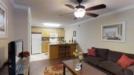This single-floor space offers an open-concept design that integrates the living room and kitchen into one cohesive, functional area. The living room is anchored by a dark, cushioned sofa accented with red throw pillows and complemented by a wooden coffee table, creating a warm and inviting atmosphere. A ceiling fan with a built-in light fixture is centrally mounted, providing both airflow and ambient lighting, while crown molding along the upper walls adds a refined architectural detail.

The kitchen features light wood cabinetry that extends to the upper walls, offering ample storage. White appliances, including a refrigerator, microwave, and range, provide a clean, classic look. A raised breakfast bar with dark granite-style countertops separates the kitchen from the living area, accompanied by two bar stools that make it ideal for casual dining. Tile flooring in the kitchen transitions to rich hardwood-style flooring in the living area, adding visual contrast. A decorative area rug with floral patterns grounds the living space, enhancing the overall warmth and character of this well-appointed home.