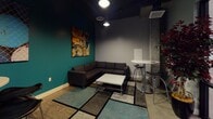 Located at 55 N Fifth St in downtown San Jose, this single-floor commercial lounge space offers a contemporary, urban aesthetic defined by a striking contrast of teal accent walls and neutral gray surfaces. The interior design balances creative expression with functional layout, making it well-suited for both social and professional gatherings.

The main lounge area features a generously sized charcoal sectional sofa arranged around a low white coffee table, anchored by a patterned area rug that adds texture and warmth to the polished concrete flooring. A large, vibrant abstract painting adorns the teal accent wall, serving as a bold focal point. Overhead, recessed and pendant lighting illuminate the space with a focused, ambient glow beneath a dark ceiling that adds an industrial character.

Adjacent to the lounge seating is a bistro-style high table paired with sleek black bar stools, offering an alternative spot for casual meetings or informal conversations. A second high table with additional bar seating is positioned nearby, accompanied by another striking large-format photograph on the teal wall.

A wall-mounted flat-screen television is visible near the entrance, which features black-framed glass double doors opening to a broader shared corridor. Exposed ductwork and an industrial ceiling treatment reinforce the space's urban, loft-inspired character throughout.