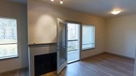 This single-floor space offers a clean, modern aesthetic with a welcoming and functional layout. The living area serves as the heart of the space, highlighted by a stylish fireplace with a tile surround and a sleek mantle that adds both warmth and character to the room. Smooth, hardwood-style flooring runs throughout, contributing to a cohesive and polished look underfoot.

Natural light fills the space generously, courtesy of a large window fitted with blinds for adjustable privacy, and a full glass-paneled door that opens directly onto a private outdoor balcony. The balcony, visible through the door, features a white railing and offers a connected indoor-outdoor living experience. The door design, with its integrated glass panel, further enhances the flow of light into the interior.

Overhead, track lighting provides focused, adjustable illumination, complementing the natural light and ensuring the space remains bright at any hour. The walls are finished in a neutral, warm tone that creates a calm and inviting ambiance throughout. The overall design is practical yet refined, making thoughtful use of space while maintaining a comfortable and airy atmosphere suited to everyday living.