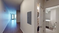 This single-floor unit showcases a thoughtfully designed open-concept layout with a distinctly modern, urban aesthetic. Warm hardwood-style flooring flows throughout the living spaces, complementing the clean white walls and lending a cohesive feel to the interior.

The kitchen is a standout feature, outfitted with a full suite of stainless steel appliances — including a refrigerator, electric range with microwave above, and dishwasher — paired with sleek, flat-front white cabinetry and a simple, functional countertop. An exposed industrial-style ductwork runs prominently overhead, adding a loft-like character to the space.

The open living area is generously sized, benefiting from large windows that invite an abundance of natural light and offer views of the surrounding urban landscape. A ceiling fan adds comfort and airflow to this bright, airy room.

The bedroom is spacious and calm, featuring sliding closet doors with built-in rod storage and continued hardwood-style flooring. The exposed ductwork carries through here as well, maintaining the industrial design language.

The bathroom is clean and contemporary, appointed with a wide vanity, ample drawer and cabinet storage, a full-width mirror, and elegant pendant lighting above. Neutral tile flooring completes the refined look.

A dedicated in-unit laundry closet with a stacked washer and dryer provides added convenience, while an accessible electrical panel is neatly integrated into the hallway wall.
