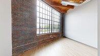 Located at 470 Market Ave SW in the heart of Grand Rapids, MI, this one-bedroom, one-bathroom loft seamlessly blends historic industrial character with modern finishes. The space is defined by soaring exposed wood beam ceilings with visible joists, original brick walls, and light hardwood flooring that runs throughout, creating a warm yet contemporary aesthetic.

The kitchen is thoughtfully designed with sleek gray cabinetry, white subway tile backsplash, stainless steel appliances — including a refrigerator, range, microwave, and dishwasher — and a clean white countertop with an undermount sink featuring matte black fixtures. A striking barn-style sliding door with a black steel grid frame adds an industrial accent at the kitchen entry.

The living area is a standout feature of the space, anchored by a dramatic floor-to-ceiling grid window wall that floods the room with natural light and frames views of the surrounding waterway and cityscape. The exposed brick wall adds raw texture and character, reinforcing the loft's industrial roots.

The bedroom offers a cozy, well-defined retreat, while the bathroom is finished with large-format gray floor tiles, white herringbone-patterned subway tile in the tub surround, and coordinating matte black hardware and fixtures throughout. The overall ambiance is urban and refined, ideal for those who appreciate architectural history paired with contemporary comfort.