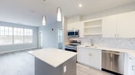 Located in East Providence, Rhode Island, this single-floor residence offers a thoughtfully designed layout with modern finishes throughout. The kitchen serves as a central highlight, featuring crisp white cabinetry, a striking marble-patterned tile backsplash, quartz countertops, and a full suite of stainless steel appliances including a range, over-the-range microwave, dishwasher, and refrigerator. A large center island with a light-colored countertop and pendant lighting above provides additional prep space and casual seating. Light wood-tone luxury vinyl plank flooring flows through the kitchen and main living areas, lending warmth and continuity to the open layout. The living and dining areas are bright and airy, benefiting from multiple windows dressed with blinds that invite natural light while maintaining privacy. The bedrooms feature soft neutral carpet and generously sized windows offering views of the surrounding neighborhood. The bathroom is well-appointed with a soaking tub and shower combination, wood-look tile flooring, and a vanity with warm-toned cabinetry. Soft blue-gray walls are consistent throughout the residence, contributing to a calm, contemporary atmosphere. The overall design balances modern aesthetics with functional living spaces.
