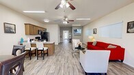 This well-appointed single-floor space offers an open-concept layout that seamlessly connects the living, dining, and kitchen areas into one cohesive, functional environment. Light-toned wood-look tile flooring runs throughout, lending a clean and modern aesthetic that ties each zone together.

The living area is generously proportioned, featuring a bold red sofa, neutral accent chairs, and a glass coffee table arranged around a wall-mounted flat-screen television. Multiple windows fitted with white horizontal blinds allow natural light to fill the room, while ceiling fans with integrated lighting ensure year-round comfort. Framed artwork adds personality to the crisp, neutral walls.

Adjacent to the living space, a dedicated dining nook accommodates a round table with seating for four, positioned beneath a window that provides additional natural light. The kitchen is fully equipped with warm wood cabinetry, granite-style countertops, a breakfast bar with seating, and a full suite of black appliances including a refrigerator, range, and over-the-range microwave. A utility sink and ample counter space complete the practical kitchen setup.

A hallway leads to an accessible bathroom finished with dark tile wainscoting, a wall-mounted sink, grab bars, and a large mirror — thoughtfully designed to accommodate a range of mobility needs. The space also features a glass-panel entry door that draws in additional light from outside.