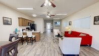 This well-appointed single-floor space offers an open-concept layout that seamlessly connects the living, dining, and kitchen areas into one cohesive, functional environment. Light-toned wood-look tile flooring runs throughout, lending a clean and modern aesthetic that ties each zone together.

The living area is generously proportioned, featuring a bold red sofa, neutral accent chairs, and a glass coffee table arranged around a wall-mounted flat-screen television. Multiple windows fitted with white horizontal blinds allow natural light to fill the room, while ceiling fans with integrated lighting ensure year-round comfort. Framed artwork adds personality to the crisp, neutral walls.

Adjacent to the living space, a dedicated dining nook accommodates a round table with seating for four, positioned beneath a window that provides additional natural light. The kitchen is fully equipped with warm wood cabinetry, granite-style countertops, a breakfast bar with seating, and a full suite of black appliances including a refrigerator, range, and over-the-range microwave. A utility sink and ample counter space complete the practical kitchen setup.

A hallway leads to an accessible bathroom finished with dark tile wainscoting, a wall-mounted sink, grab bars, and a large mirror — thoughtfully designed to accommodate a range of mobility needs. The space also features a glass-panel entry door that draws in additional light from outside.
