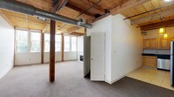 Unit 209 offers a distinctive loft-style living experience defined by its industrial-meets-rustic character. Throughout the unit, warm wood plank ceilings with exposed timber beams and original structural columns create a strong architectural identity, complemented by white walls and neutral carpet flooring in the living and bedroom areas.

The expansive main living area features exposed ductwork, a ceiling fan, and rows of large multi-pane windows that flood the space with natural light and provide open views of the surrounding neighborhood. The kitchen is equipped with warm-toned wood cabinetry, dark granite countertops, tile flooring, and a full suite of stainless steel appliances, including a refrigerator, range, microwave, and dishwasher, all accented by pendant lighting beneath the beamed ceiling.

Both bedrooms are generously proportioned, carpeted, and bathed in natural light from banks of large windows. Each bedroom includes ceiling fans and access to closet space. The two full bathrooms are finished with dark vanity cabinets, white countertops, tub/shower combinations, tile flooring, and lighted mirrors, maintaining a clean, functional aesthetic consistent throughout the unit.