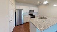 This single-floor residence is designed with a clean, modern aesthetic and a thoughtful layout that balances functionality with comfort. The open-concept living and kitchen area serves as the heart of the home, featuring rich, dark hardwood-style flooring that extends throughout the main living spaces. A ceiling fan overhead provides both airflow and ambient lighting, while multiple windows dressed with blinds allow natural light to filter in, creating a bright and welcoming atmosphere.

The kitchen is a standout feature, outfitted with crisp white shaker-style cabinetry that extends to the ceiling for generous storage. Granite countertops with a subtle speckled pattern run along the counters and across a central island that includes a built-in sink. Stainless steel appliances, including a refrigerator, range, and over-the-range microwave, add a sleek, contemporary touch. A tile backsplash completes the polished look.

The bedroom offers a comfortable retreat with plush wall-to-wall carpeting, a ceiling fan with integrated lighting, and a window that brings in natural light. A walk-in closet with built-in shelving provides ample storage, and a panel door leads to the hallway.

The bathroom continues the refined finishes seen throughout the home, with granite-topped vanity cabinetry, wood-style flooring, and a clean, functional layout. Overall, the space presents a move-in-ready environment with consistent, quality finishes.