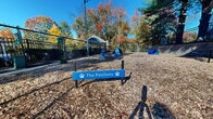 The Pavilions is a dedicated outdoor dog park located at 465 Buckland Hills Dr in Manchester, Connecticut. The space is entirely open-air and single-level, featuring a generously sized, wood chip-covered play area that provides a comfortable, natural surface underfoot for pets. The park is fully enclosed by sturdy black iron fencing, ensuring a safe and secure environment for dogs to roam freely.

A covered gazebo-style shelter anchors one end of the park, offering shade and a gathering point for visitors. Playful, dog-themed features are distributed throughout the space, including decorative fire hydrants in both blue and red, as well as a small pet tunnel for enrichment and play. Signage throughout the area is branded in blue with paw print motifs, reinforcing the park's welcoming, pet-focused identity.

The park is bordered by tiered stone retaining walls that add structural definition and a landscaped aesthetic to the perimeter. Mature deciduous and evergreen trees surround the fenced boundary, providing natural shade and vibrant seasonal color, particularly vivid during the autumn months. Overhead lighting is available via tall pole-mounted fixtures, extending usability into the evening. The overall atmosphere is open, bright, and well-maintained, offering a pleasant outdoor experience for both pets and their owners.