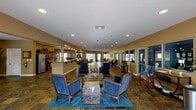 This single-story community clubhouse offers a thoughtfully designed open-plan layout that seamlessly connects multiple functional areas under one roof. The main lounge serves as the centerpiece of the space, featuring rich slate tile flooring in warm earthy tones, recessed lighting, and comfortable blue upholstered seating arranged around a central coffee table atop a vibrant blue area rug. The open design allows natural light to filter in through large windows and glass doors along the perimeter, creating a bright and welcoming atmosphere throughout.

Adjacent to the lounge is a fully equipped kitchen, outfitted with rich wood cabinetry, granite countertops, stainless steel appliances including a gas range, dishwasher, and refrigerator, making it well-suited for community events and gatherings. A casual dining area nearby features a round wood table with wicker chairs and framed artwork lining the walls, providing an inviting space for smaller groups.

The facility also includes a dedicated office area with an interior service window, private offices accessible via glass-panel doors, and a small children's activity corner with a pint-sized table and stools. Decorative potted plants and curated artwork throughout add warmth and character, rounding out this versatile and functional community space.