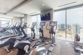 This 1,670-square-foot fitness center is a comprehensively outfitted facility designed to support a wide range of workout routines. The space is organized into distinct functional zones, beginning with an extensive cardio area lined with treadmills, ellipticals, stair climbers, and stationary bikes—each equipped with individual digital screens for an engaging workout experience. A dedicated strength training section features a multi-functional cable and weight machine system alongside benches and free weights. A separate free weights and functional training area offers barbells, weight plates, medicine balls, resistance bands, and exercise mats, all set on durable rubber flooring ideal for dynamic movements. Large wall mirrors throughout enhance the sense of space and support form monitoring. The facility is finished with a drop ceiling featuring recessed lighting, and patterned carpet flooring in the cardio zone. Floor-to-ceiling windows wrap around multiple sides of the space, flooding the interior with natural light and offering sweeping panoramic views of the surrounding cityscape—making every workout session as visually inspiring as it is physically rewarding.