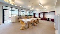 Located at 1340 S Hill St in the heart of Downtown Los Angeles, this single-floor commercial space offers a well-appointed layout comprising a private conference room and a vibrant communal dining area. The conference room is bright and functional, featuring floor-to-ceiling black-framed windows that frame views of the surrounding cityscape and flood the room with natural light. A sculptural gold loop chandelier serves as a striking focal point above a sleek light wood conference table surrounded by modern cantilever chairs. A wall-mounted display screen adds to the room's professional utility, making it ideal for meetings and presentations. The teal patterned carpet adds a bold pop of color. The dining area exudes a warm, inviting ambiance with a large communal table surrounded by woven rattan chairs in golden tones. Elegant pendant chandeliers with layered white shades cast a soft glow over the space. Along one wall, a planter-topped bar counter with high stools offers additional seating beside the windows. Cozy high-back booth seating lines one side, while a row of semi-private upholstered pod chairs — adorned with decorative woven cushions — lines the opposite wall beneath a curated arrangement of decorative juju hat wall art. Rich burgundy accent alcoves add depth and warmth, completing this thoughtfully curated, multifunctional space.