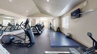Located at 353 Dalton St in Medford, Oregon, this fitness center offers a comprehensive, single-level exercise facility designed to support a wide range of workout routines. The space is characterized by its vaulted, angled ceilings that lend an open, airy feel, complemented by recessed lighting and natural light streaming in through multiple windows fitted with blinds.

The main exercise area features dark rubber flooring throughout, providing both durability and safety underfoot. A row of treadmills lines one side of the room, accompanied by elliptical trainers and a recumbent bike, catering to a variety of cardiovascular training preferences. A wall-mounted flat-screen TV is positioned for easy viewing during workouts.

A dedicated stretching and floor exercise zone is defined by a large blue gymnastics mat, alongside a stability ball and a rowing machine, offering ample space for functional and flexibility training. A separate strength training alcove houses a multi-station cable machine, a weight bench, and a rack of dumbbells, providing a well-rounded resistance training setup.

Additional amenities include a sanitation station stocked with cleaning supplies, posted facility rules, and a wall-mounted clock. The space benefits from a mirror along one wall, enhancing the sense of spaciousness. The overall ambiance is clean, bright, and purpose-built for effective fitness use.