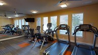 This fitness center is designed to provide a comprehensive and motivating workout environment. The space is outfitted with a variety of cardio equipment, including treadmills, an elliptical trainer, and a stationary bike, with notable Cybex-branded machines among the offerings. Strength and resistance equipment is also present, ensuring a well-rounded exercise experience. The warm, golden-yellow walls create an energizing ambiance, complemented by wood-style flooring accented with orange fitness mat sections that add both function and visual appeal. Large windows with horizontal blinds line the walls, inviting ample natural light and offering views of the surrounding greenery, contributing to an open and refreshing atmosphere. Mirrored walls on one side visually expand the space and allow users to monitor their form during workouts. A wall-mounted flat-screen television provides entertainment, and ceiling-mounted light fixtures ensure the room is well-lit at all times. A ceiling fan aids in ventilation and comfort during intense exercise sessions. The layout is thoughtfully arranged to allow ease of movement between equipment, making this fitness center both practical and inviting for daily use.
