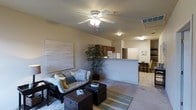 This single-floor residence offers an open-concept layout that promotes a natural flow between the living, kitchen, and dining areas. The living room is a comfortable and inviting space, featuring plush carpeting, a ceiling fan with integrated lighting, and room for a sofa, coffee table, and additional seating. A textured wall canvas and a potted indoor plant add personality and a touch of nature to the space. The kitchen is positioned just beyond a half-wall peninsula that serves as a natural divider, providing both functionality and an open sightline throughout the common areas. The kitchen features warm wood-toned cabinetry and is equipped with standard appliances, including a refrigerator and microwave. Adjacent to the kitchen, the dining area offers a cozy nook with a small table and chairs, ideal for everyday meals. A doorway from the dining area leads further into the residence, suggesting additional private rooms beyond. The overall ambiance is bright and neutral, with warm beige tones on the walls and light-colored surfaces that contribute to an airy and livable atmosphere throughout.
