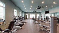 The Marq's fitness center is a generously sized, single-floor facility designed to accommodate a comprehensive range of workout needs. The space is finished in a cool blue-gray palette, lending a clean and focused atmosphere throughout. Rubber flooring in a speckled finish covers the entire area, providing durability and comfort underfoot.

The cardio zone features a long row of TRUE-brand treadmills, ellipticals, and stationary bikes, including upright and recumbent models equipped with individual display screens. Multiple large windows line the perimeter walls, allowing substantial natural light to fill the space while offering views of the surrounding community.

The strength training area is outfitted with a variety of cable and selectorized machines, free weights, a dumbbell rack, and adjustable benches, providing options for both resistance and functional training. Full-length mirrors along one wall visually expand the space and allow users to monitor their form. Mounted flat-screen televisions are positioned throughout for entertainment during workouts.

Overhead, large ceiling fans provide consistent air circulation, complemented by recessed lighting that ensures even illumination. A built-in storage area with open cubbies and navy cabinetry adds organizational convenience. The overall layout is open and uncluttered, supporting an effective and comfortable workout environment.