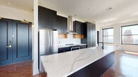 This thoughtfully designed single-floor space showcases a seamless blend of contemporary aesthetics and functional living. The centerpiece of the home is a dramatic chef's kitchen, anchored by an expansive waterfall island finished in white marble-veined stone. Floor-to-ceiling matte dark cabinetry with warm brass hardware creates a bold contrast against a classic white subway tile backsplash, while open natural wood shelving adds an organic touch. Stainless steel appliances, including a full-size refrigerator and range with a sleek wall-mounted ventilation hood, complete the culinary suite. A matte black faucet over an integrated sink furthers the kitchen's refined, high-contrast design language. Throughout the main living areas, rich herringbone-patterned hardwood flooring adds texture and warmth underfoot, complementing the neutral tones of the walls. Bedrooms are generously proportioned, each benefiting from large windows that invite natural light and offer elevated city views. Interior doors are finished in a deep matte black with brass hardware, maintaining design continuity across the space. A powder room features vertically stacked tile detailing, light wood flooring, and a pedestal sink. A utility area with dark slate tile flooring and a window overlooking city rooftops rounds out this well-appointed home.