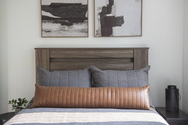 Contemporary bedroom with a stylish wooden headboard, cozy gray and brown pillows, and modern abstract wall art. - Grace Lane Apartments