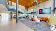 This modern commercial facility makes an immediate impression with its grand, double-height atrium lobby, anchored by a striking glass-railed staircase, soaring wood-slat ceilings, and bold cylindrical columns. The main atrium flows seamlessly into multiple seating zones furnished with leather sofas, accent chairs in warm cognac and sage tones, and patterned area rugs — all bathed in natural light streaming through floor-to-ceiling glazed walls that offer views of the surrounding landscape. A central corridor connects the atrium to the broader facility, lined with built-in planters featuring ornamental grasses and sculptural accents that add a biophilic warmth to the refined interiors. The open-plan workspace area is defined by a slatted wood ceiling, warm walnut desking, and sleek black task chairs, complemented by ample natural light and wall-mounted displays. A café lounge features a tiled counter bar with pendant lighting, large windows overlooking palm trees, and mixed seating arrangements ideal for informal gatherings. Additional lounge rooms offer more intimate settings with leather seating and outdoor access. Large-format porcelain tile flooring runs throughout, unifying the space with a clean, professional aesthetic.