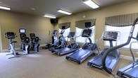 This fitness center offers a clean and functional environment designed to support a well-rounded cardio workout. The room is finished with warm beige walls and neutral carpeted flooring, creating a welcoming atmosphere conducive to exercise. Overhead fluorescent lighting ensures the space is well-illuminated throughout, while multiple windows dressed with valance treatments allow natural light to filter in, adding brightness and ventilation to the room.

The equipment lineup includes multiple Star Trac treadmills, elliptical cross-trainers, and upright stationary bikes, each unit featuring individual digital display consoles for tracking workout metrics. A wall-mounted television is positioned for easy viewing during workouts, adding an entertainment element to the exercise experience.

The layout is thoughtfully arranged to provide ample room between machines, allowing for comfortable and safe use of each station. A glass-paneled door provides access to the space, contributing additional light and a sense of openness. The overall design strikes a balance between practicality and comfort, making this fitness center a convenient and inviting option for daily exercise.