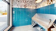 Located at 414 Front St in Santa Cruz, California, this purpose-built pet grooming room is a compact yet fully functional single-room space designed specifically for pet care. The room's most striking design element is its vibrant, glossy blue subway tile that lines the walls from floor to mid-height, creating a clean, water-resistant surface well-suited for its grooming function. Above the tile, a playful black-and-white illustrated mural wraps around the upper walls, depicting a whimsical crowd of cartoon dogs and cats, giving the space an inviting and lighthearted character. A flush-mounted circular ceiling light provides even, practical illumination throughout the room. The centerpiece of the space is a professional-grade stainless steel pet wash tub, complete with an integrated ramp and steps for easy pet access, a handheld sprayer, and a floor drain nearby to manage water efficiently. A built-in wooden storage unit with drawers offers convenient organization for grooming supplies. A glass door and large window along one wall allow natural light to enter and provide a view of the outdoor pedestrian plaza beyond, connecting the interior to its lively urban surroundings. The polished concrete floor completes the practical, easy-to-clean aesthetic of this well-appointed grooming station.