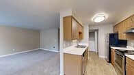 This single-floor, 2-bedroom, 1-bathroom home offers a practical and comfortable layout across a well-utilized footprint. The open-concept kitchen flows seamlessly into the living area, which features neutral-toned carpet and a window that allows natural light to fill the space. The kitchen is efficiently designed with warm wood-toned cabinetry, white countertops, a double-basin stainless steel sink, and black and stainless steel appliances including an electric range and dishwasher. A chandelier overhead adds a subtle decorative touch to the dining area adjacent to the kitchen. In-unit washer and dryer hookups are conveniently accessible from the kitchen corridor. Both bedrooms are carpeted and finished in soft neutral tones, with the primary bedroom offering a comfortable amount of space. Each bedroom includes closet storage, with built-in hanging rods providing organized storage solutions. The bathroom is well-appointed with wood-toned cabinetry, a single vanity with a large mirror, and a full tub-shower combo. Light wood-look tile flooring completes the bathroom's clean, functional aesthetic. A hallway connects the main living spaces, and the overall palette of warm beiges and taupes gives the home a cohesive, inviting character throughout.