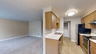 This single-floor, 2-bedroom, 1-bathroom home offers a practical and comfortable layout across a well-utilized footprint. The open-concept kitchen flows seamlessly into the living area, which features neutral-toned carpet and a window that allows natural light to fill the space. The kitchen is efficiently designed with warm wood-toned cabinetry, white countertops, a double-basin stainless steel sink, and black and stainless steel appliances including an electric range and dishwasher. A chandelier overhead adds a subtle decorative touch to the dining area adjacent to the kitchen. In-unit washer and dryer hookups are conveniently accessible from the kitchen corridor. Both bedrooms are carpeted and finished in soft neutral tones, with the primary bedroom offering a comfortable amount of space. Each bedroom includes closet storage, with built-in hanging rods providing organized storage solutions. The bathroom is well-appointed with wood-toned cabinetry, a single vanity with a large mirror, and a full tub-shower combo. Light wood-look tile flooring completes the bathroom's clean, functional aesthetic. A hallway connects the main living spaces, and the overall palette of warm beiges and taupes gives the home a cohesive, inviting character throughout.
