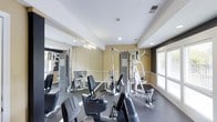 This dedicated fitness center is thoughtfully designed to support a well-rounded workout experience. The space is outfitted with a selection of strength training machines and cardio equipment, providing users with diverse exercise options. Large mirrors line one wall, enhancing the sense of space while allowing users to monitor their form during workouts. The flooring is a durable, dark rubberized material well-suited for a high-traffic exercise environment. Recessed lighting in the white ceiling provides even, functional illumination throughout the room, complemented by natural light streaming in through windows along the exterior wall. The walls are painted in a warm neutral tone with a contrasting darker accent, giving the space a clean and focused aesthetic. A chandelier-style light fixture adds a subtle decorative touch near the mirrored wall. The overall layout is open and practical, with equipment arranged to allow comfortable movement between stations. This single-floor fitness facility offers a functional and well-maintained environment suitable for a variety of fitness routines.