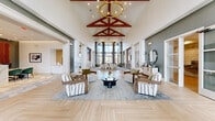 This expansive single-floor clubhouse offers a thoughtfully designed interior that blends modern elegance with warm, inviting architectural details. The grand main hall commands immediate attention with its dramatic vaulted ceiling accented by rich red-toned timber trusses and a striking brass chandelier, all above a beautifully laid herringbone hardwood floor. Tall floor-to-ceiling windows flood the space with natural light and offer views of the surrounding exterior. The living area features curated seating arrangements with patterned armchairs, layered area rugs, and accent consoles set against deep teal feature walls, creating intimate conversation zones within the open layout. A dedicated reception area showcases a sleek marble-topped desk, teal velvet chairs, and an ornate copper lattice feature wall as a bold focal point. The dining zone is anchored by white quartz countertops, exposed wood ceiling beams, and rows of black bistro-style bar seating beneath oversized pendant lights, with large windows framing outdoor views. The kitchen is outfitted with stainless steel appliances, a generous island, and seamlessly connects to the dining space. Throughout, curated artwork, accent niches in contrasting colors, and high-end finishes contribute to a sophisticated yet welcoming ambiance.