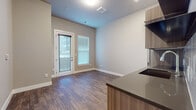 This well-appointed single-floor unit showcases a thoughtfully designed open-concept layout that seamlessly connects the kitchen and living areas. Rich, dark hardwood-style flooring runs throughout the entire space, lending warmth and visual continuity to every room.

The kitchen is a standout feature, outfitted with sleek wood-grain cabinetry, dark countertops, and a full suite of stainless steel appliances including a refrigerator, dishwasher, and sink. A white subway tile backsplash adds a clean, modern touch, while under-cabinet lighting enhances the workspace.

The living and dining area is open and airy, benefiting from natural light streaming through a large glass door and an adjacent window fitted with blinds. The glass door provides direct access to an outdoor space, creating a seamless indoor-outdoor connection. Neutral gray walls complement the flooring and offer a versatile backdrop throughout.

A dedicated wall in the living area features multiple electrical outlets and a programmable thermostat, reflecting the unit's practical, tech-ready design. The bathroom is finished with neutral tones, wood-style tile flooring, and includes a combination tub and shower with a grab bar, toilet, and clean white fixtures. Overall, the space presents a modern, functional, and cohesive aesthetic.