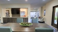 This single-story space showcases an open-concept layout that seamlessly connects the dining and living areas, creating a cohesive and functional environment. The dining area features a substantial rustic wood table paired with soft teal upholstered chairs, centered by a decorative succulent arrangement that adds a natural, organic touch.

A partial partition wall with a built-in media console anchors the space, finished in rich, dark wood paneling that provides a warm contrast to the light cream-toned walls. The built-in unit houses a large flat-screen television and is topped with a white countertop surface, offering both functionality and visual interest.

The living area beyond features an arrangement of contemporary upholstered chairs in neutral tones, creating an inviting seating space. Multiple windows fitted with horizontal blinds allow for controlled natural light throughout, maintaining a bright yet comfortable atmosphere. Recessed lighting complements the natural light, ensuring even illumination across the space. The overall design balances rustic warmth with modern, clean-lined furnishings for a refined yet approachable aesthetic.