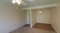 This single-floor residence offers a straightforward and functional layout designed for comfortable everyday living. The main living area is finished with wall-to-wall carpeting, providing a warm and soft underfoot feel, while a ceiling fan with a light fixture overhead ensures year-round comfort and adequate illumination. Vertical paneling along one wall adds a subtle textural detail to the space. A structural beam subtly defines the transition between the living area and the adjacent dining or secondary space, which features hardwood-style flooring that adds a clean, polished contrast to the carpeted zone. A pendant light hangs in this adjoining area, offering focused, ambient lighting. Chair rail molding lines the walls of the dining space, contributing a classic architectural touch. A white interior door provides access to additional areas of the home, hinting at a practical and connected floor plan. The overall aesthetic is clean and neutral, with light-toned walls throughout that create a bright, open atmosphere. The space presents a solid foundation well-suited for personalization and customization to suit a variety of living styles.