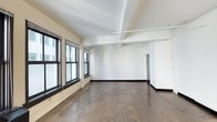 This single-floor space offers a clean, open layout with a minimalist aesthetic that lends itself to a wide range of uses. The interior is characterized by bright white walls that reflect natural light streaming in through a row of large, dark-framed windows along one side, creating a well-lit and airy atmosphere. The flooring consists of large-format dark stone-toned tiles, providing a durable and neutral foundation throughout the space. Overhead, the ceiling features exposed structural beams and utility conduit runs, lending an industrial character while maintaining a sense of height and openness. Recessed track lighting is integrated into the ceiling, offering practical and adjustable illumination. Toward the rear of the space, a doorway leads to an additional area or utility room, adding functional versatility. The open floor plan allows for flexible arrangement and configuration, making the space well-suited for office, studio, retail, or creative workspace applications. Overall, the combination of natural light, industrial detailing, and a neutral palette creates a straightforward and adaptable environment.