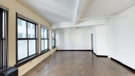 This single-floor space offers a clean, open layout with a minimalist aesthetic that lends itself to a wide range of uses. The interior is characterized by bright white walls that reflect natural light streaming in through a row of large, dark-framed windows along one side, creating a well-lit and airy atmosphere. The flooring consists of large-format dark stone-toned tiles, providing a durable and neutral foundation throughout the space. Overhead, the ceiling features exposed structural beams and utility conduit runs, lending an industrial character while maintaining a sense of height and openness. Recessed track lighting is integrated into the ceiling, offering practical and adjustable illumination. Toward the rear of the space, a doorway leads to an additional area or utility room, adding functional versatility. The open floor plan allows for flexible arrangement and configuration, making the space well-suited for office, studio, retail, or creative workspace applications. Overall, the combination of natural light, industrial detailing, and a neutral palette creates a straightforward and adaptable environment.
